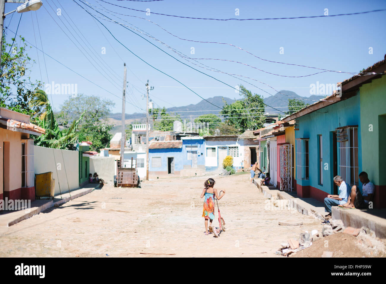 In a poor neighbourhood of Trinidad in Cuba, a girl pushes her scooter down the unpaved street lined by colourful houses Stock Photo