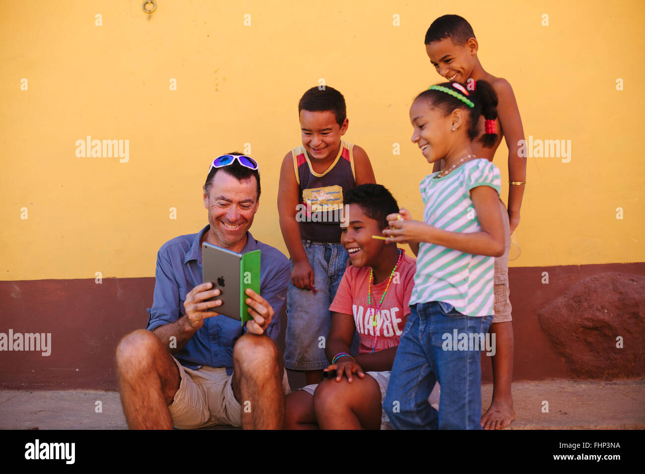 On a street in Trinidad, Cuba, a group of kids are gathered around a western male tourist and are amused by his ipad. Stock Photo
