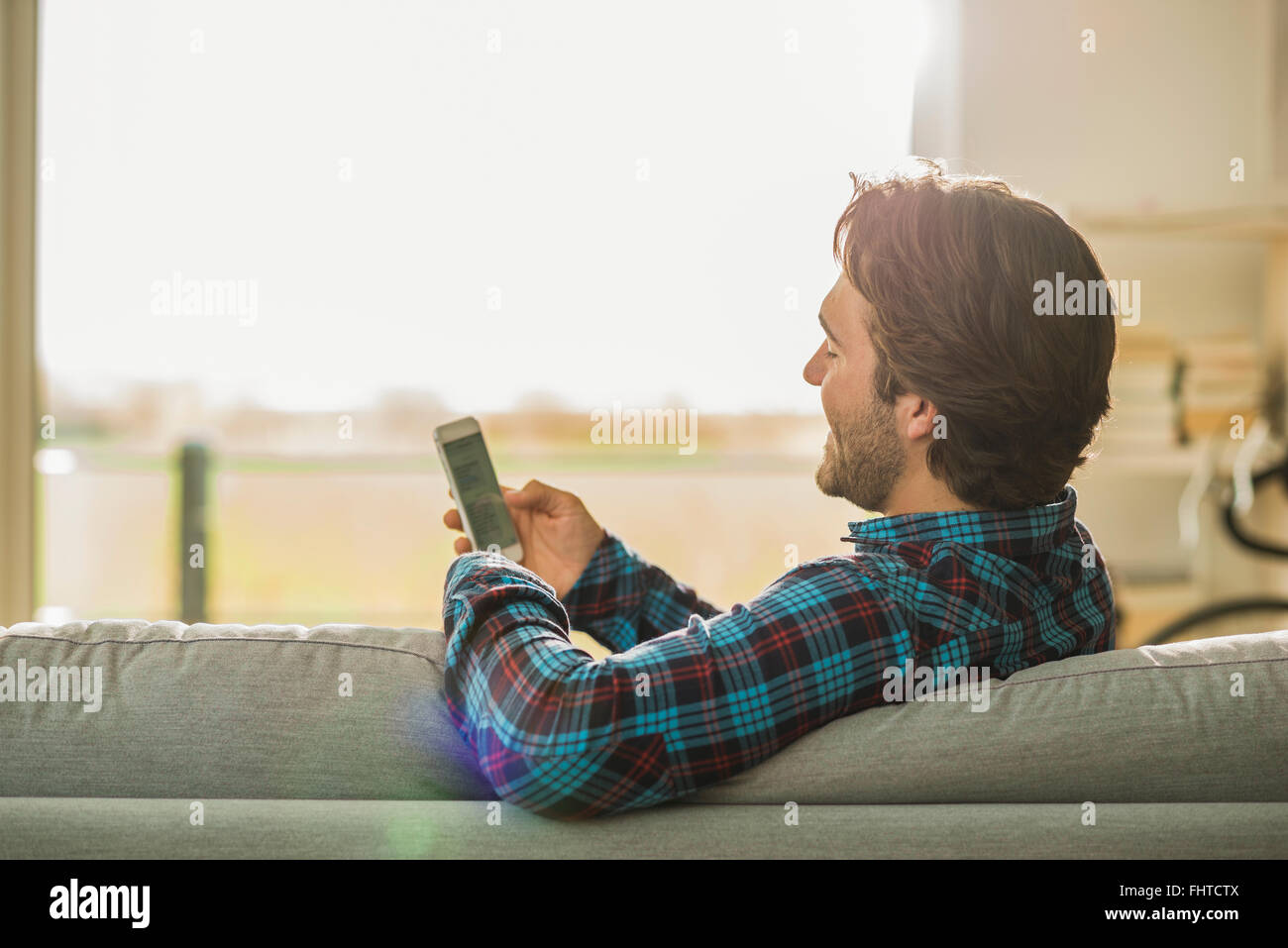 Smiling young man leaning on backrest of the couch looking at his smartphone Stock Photo