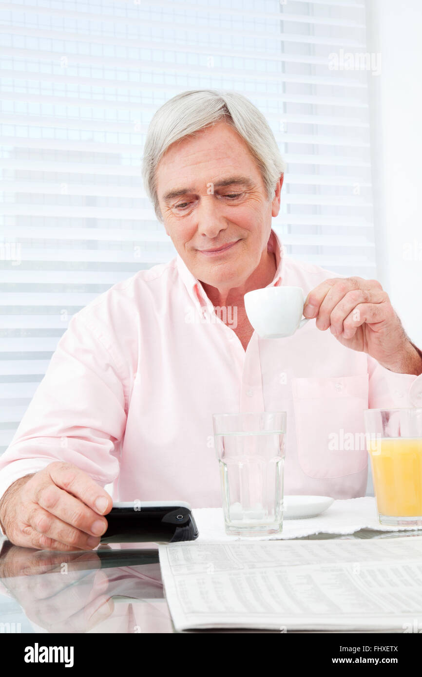 Senior man with coffee looking at smartphone at breakfast table Stock Photo