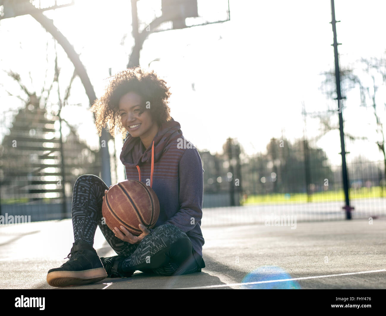 Portrait of smiling young woman with basketball sitting on a playing field Stock Photo
