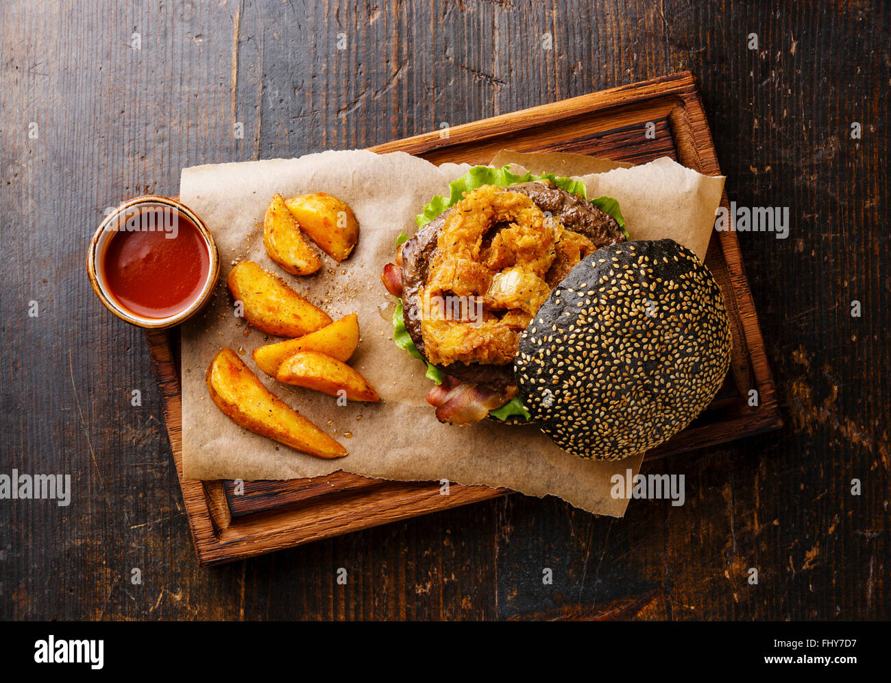 Black burger with sesame seed bun meat bacon onion rings fries and potato wedges on dark wooden background Stock Photo