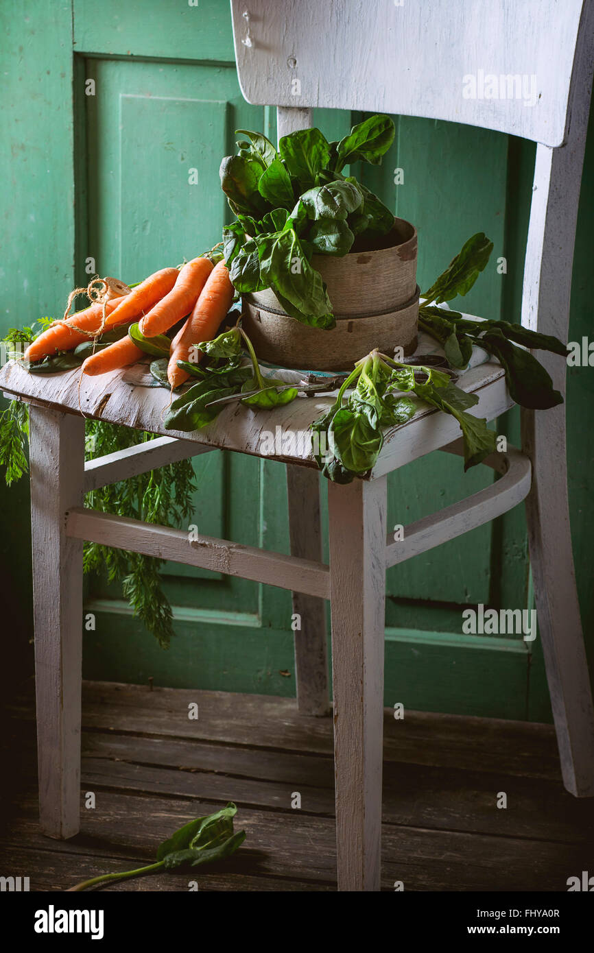 Bunch of fresh spinach and carrots on old white wooden chair with green wooden wall at background. Stock Photo