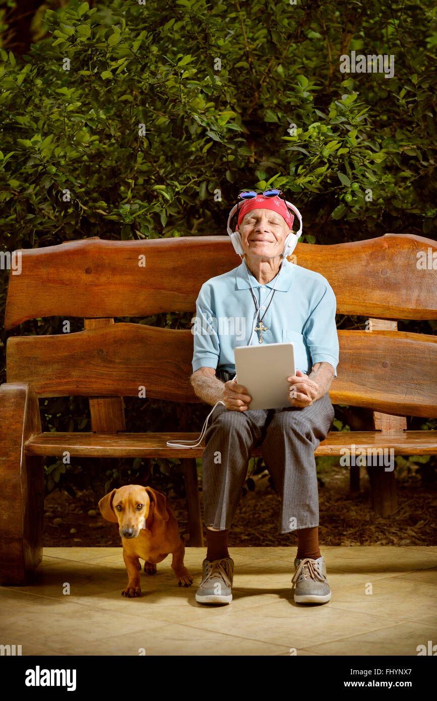 MODEL RELEASED. Senior man sitting on a bench listening to music with his dog. Stock Photo