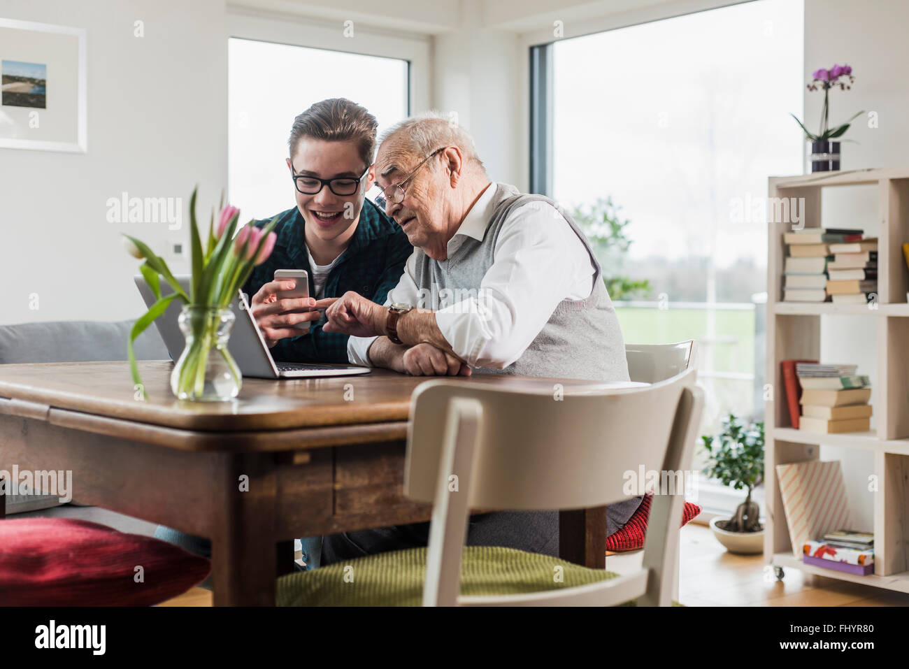 Senior man and his grandson sitting at table in the living room with laptop and smartphone Stock Photo