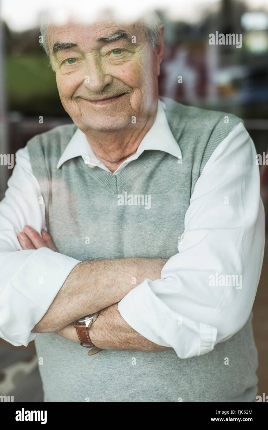 Portrait of smiling senior man with crossed arms looking through window Stock Photo