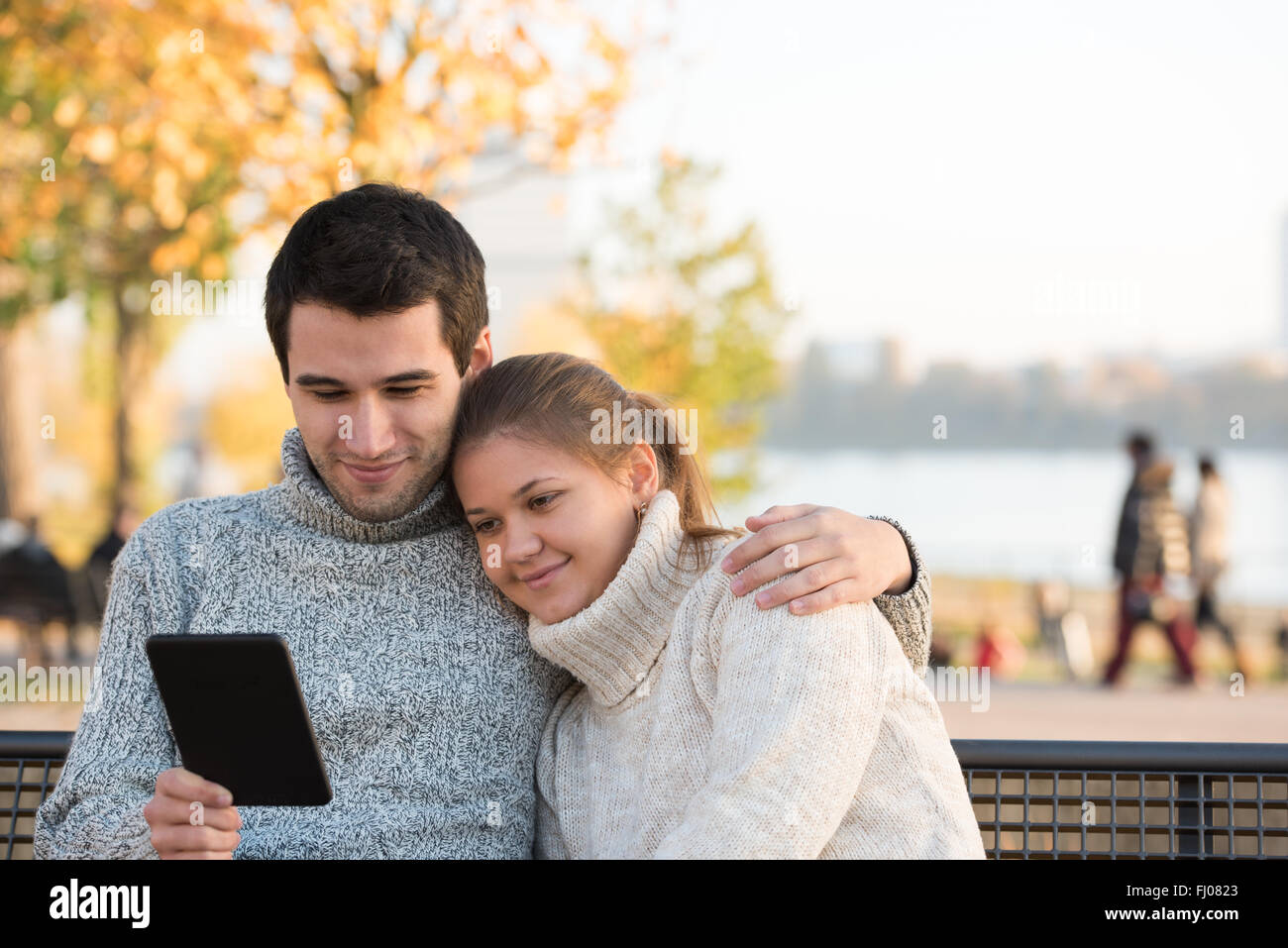 young couple in autumn park sitting on the bench, reading ebook Stock Photo