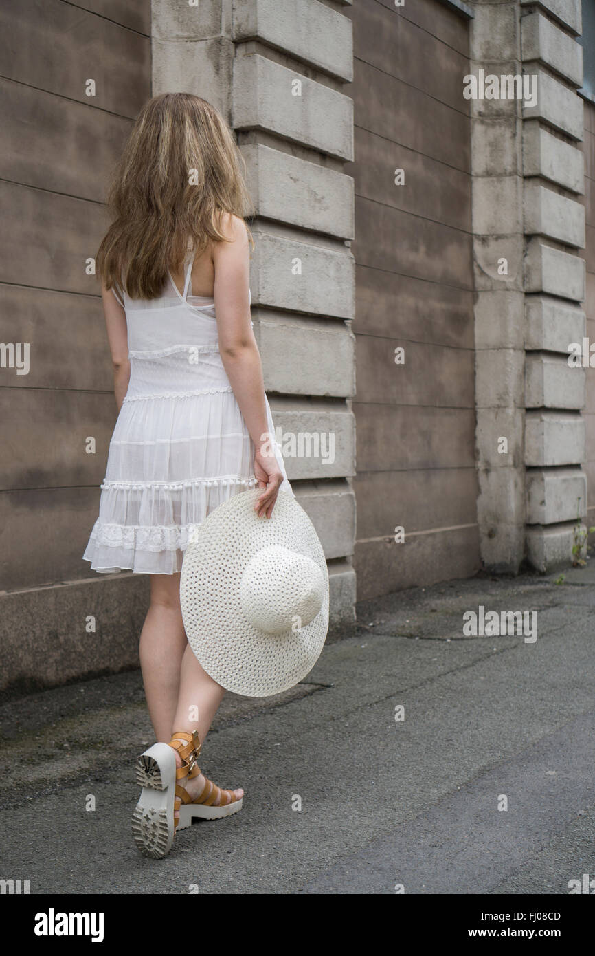 Woman wearing a vintage white dress and holding a hat walking in the street Stock Photo