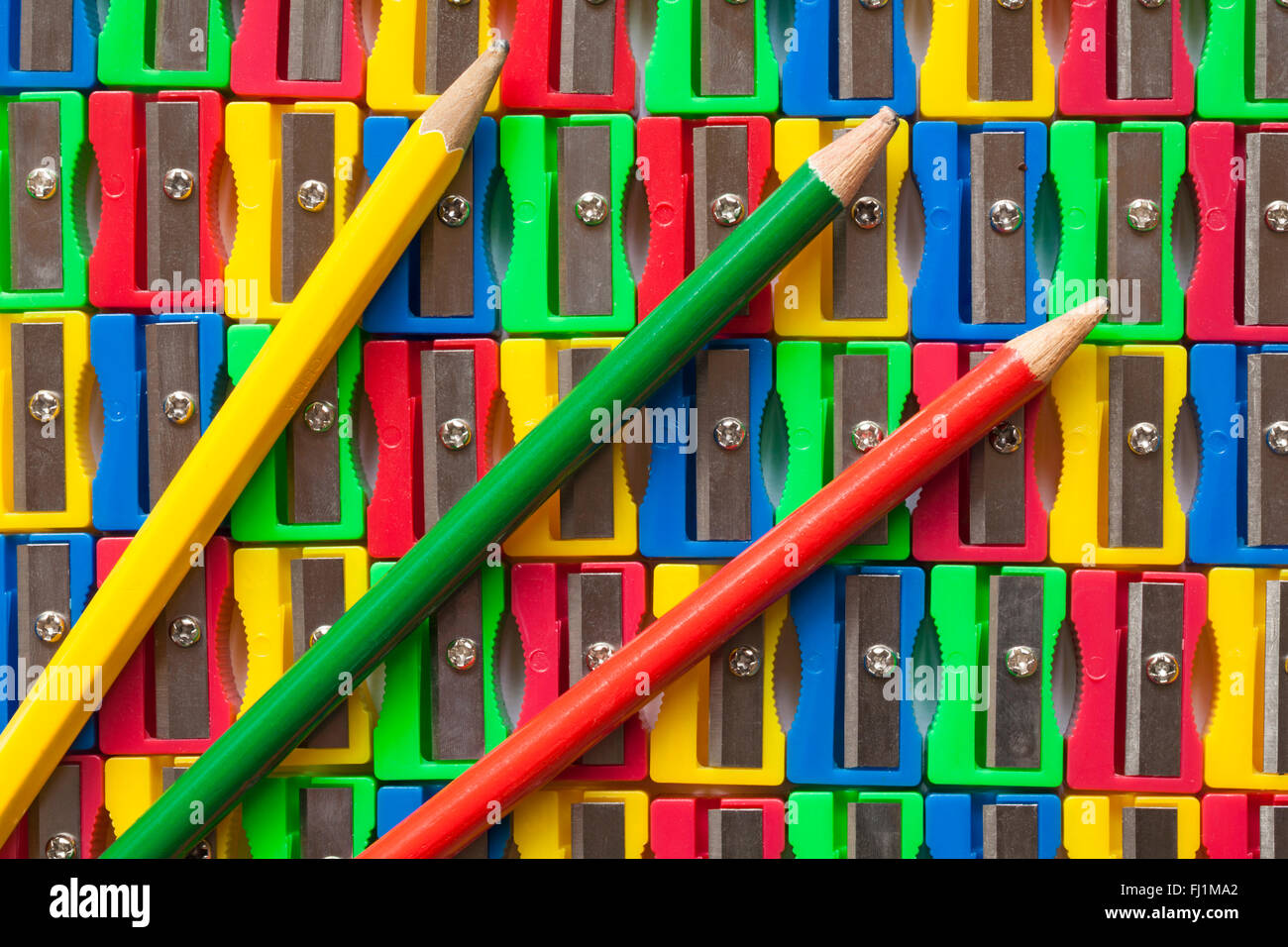 Rows of colourful red, yellow, green and blue plastic pencil sharpeners with yellow green and red pencils that need sharpening Stock Photo