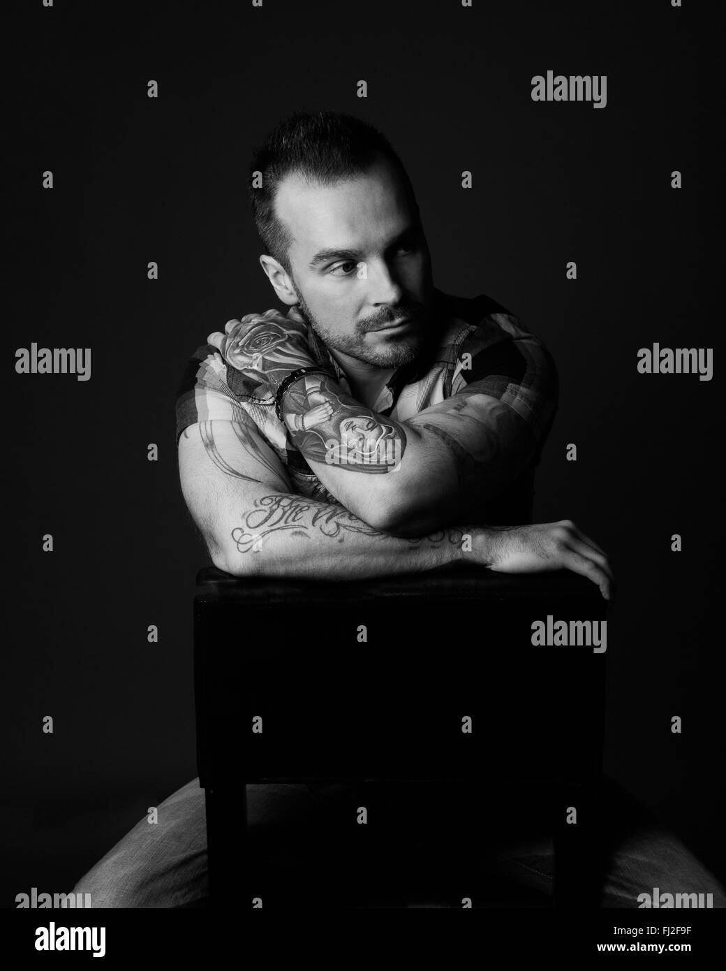 Pensive man with beard sitting on stool looking away with short sleeved shirt and tattoos in a black and white low-key setting Stock Photo