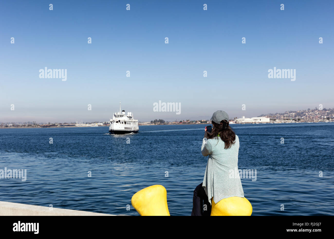 Back view of a woman taking photos with cell phone of San Diego bay in Southern California while sitting down at pier. Stock Photo