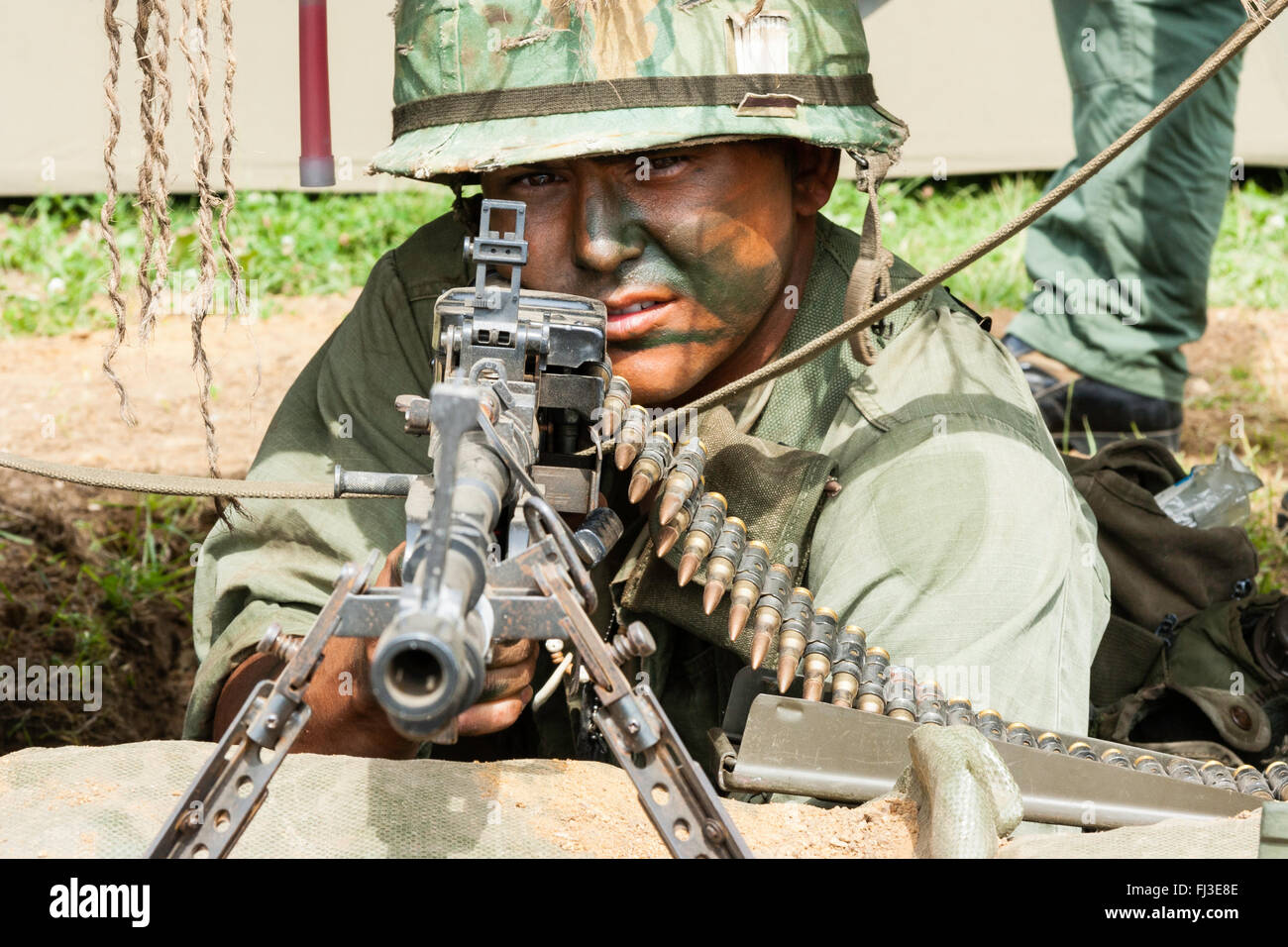 Vietnam war re-enactment. Close up. American soldier with camouflage paint on face, aiming M60 machine gun directly at viewer. Eye-contact. Stock Photo