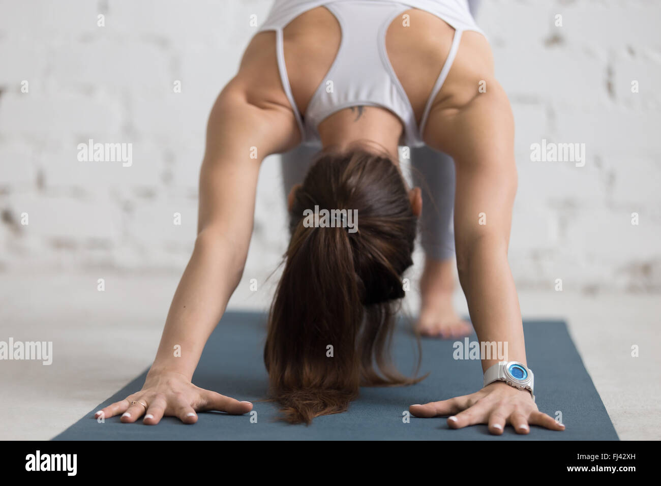 Beautiful young woman working out indoors, doing yoga exercise with sports smart watch, downward facing dog pose, adho mukha Stock Photo