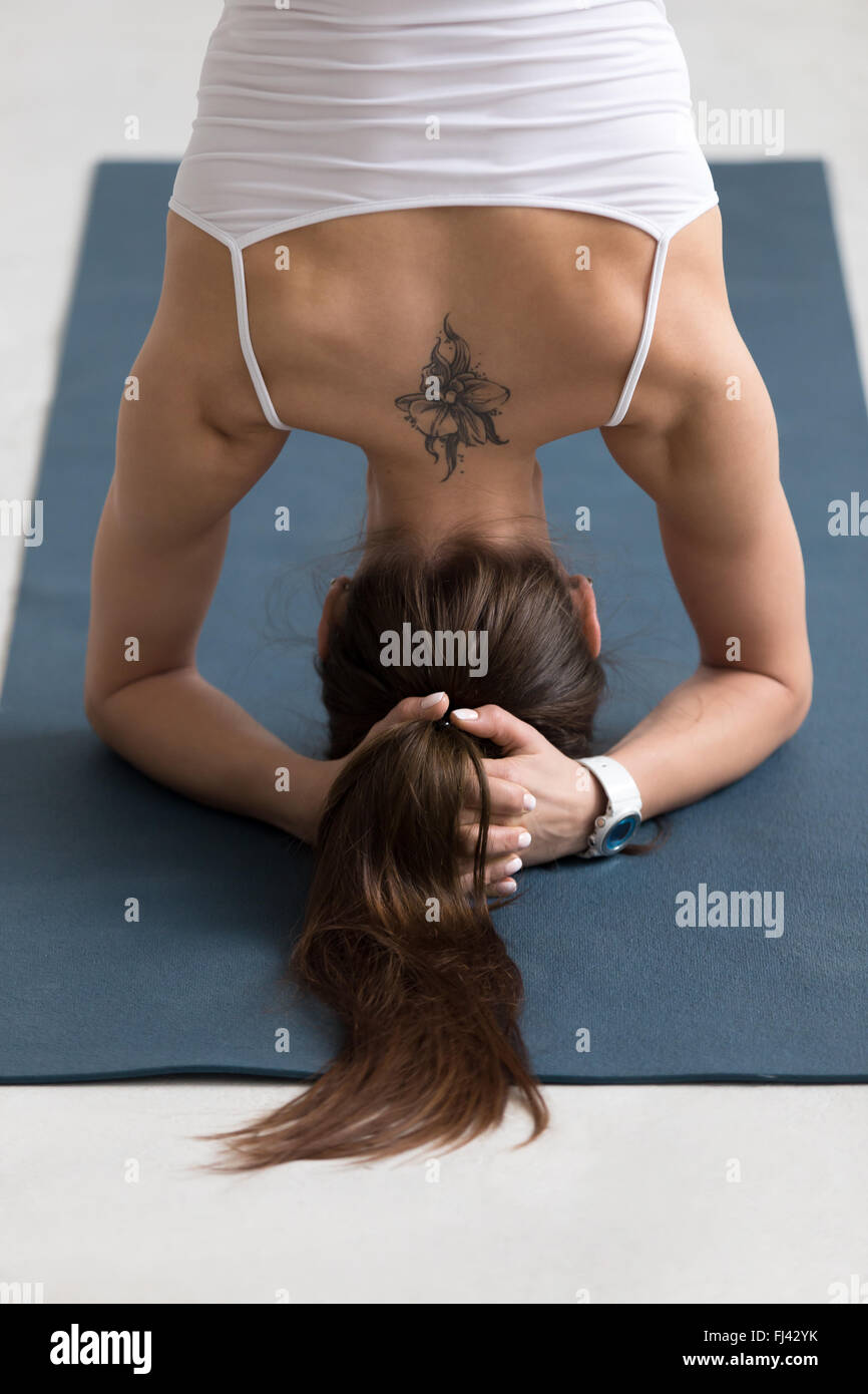 Beautiful young woman with ponytail and flower tattoo on her back working out indoors, doing yoga exercise on blue mat Stock Photo