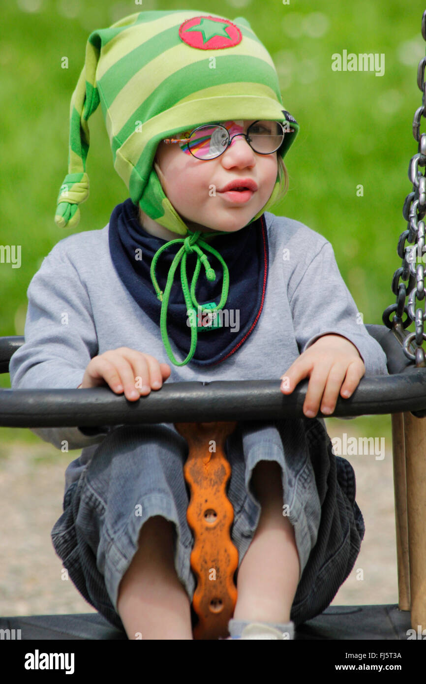 little girl with eye patch sitting amused in a swing, Germany Stock Photo