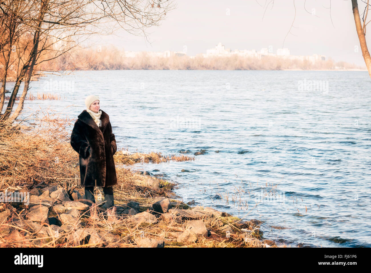 A mature woman wearing a warm fur coat and a woolen cap stays close to the Dnieper river in Kiev, Ukraine, during winter Stock Photo