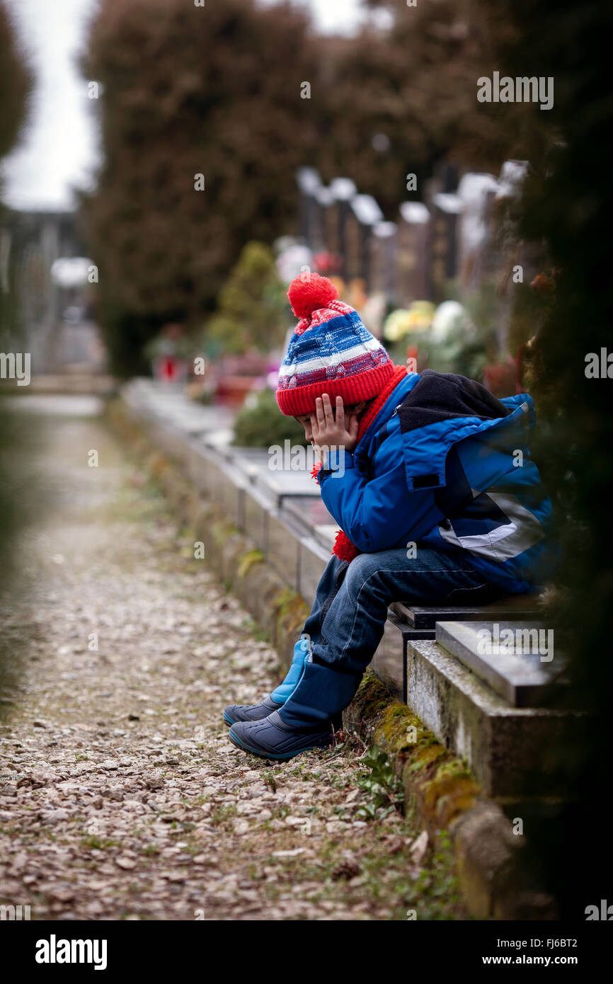 Sad little boy, sitting on a grave in a cemetery, feeling sad and ...