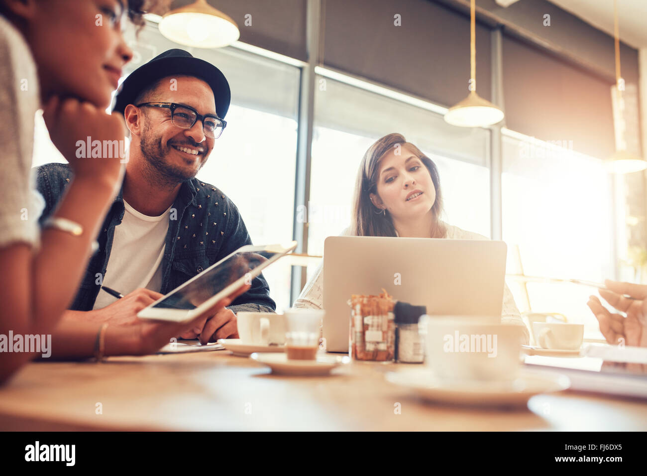 Portrait of happy young man sitting and talking with friends at a cafe. Young people at a coffee shop with digital tablet and la Stock Photo