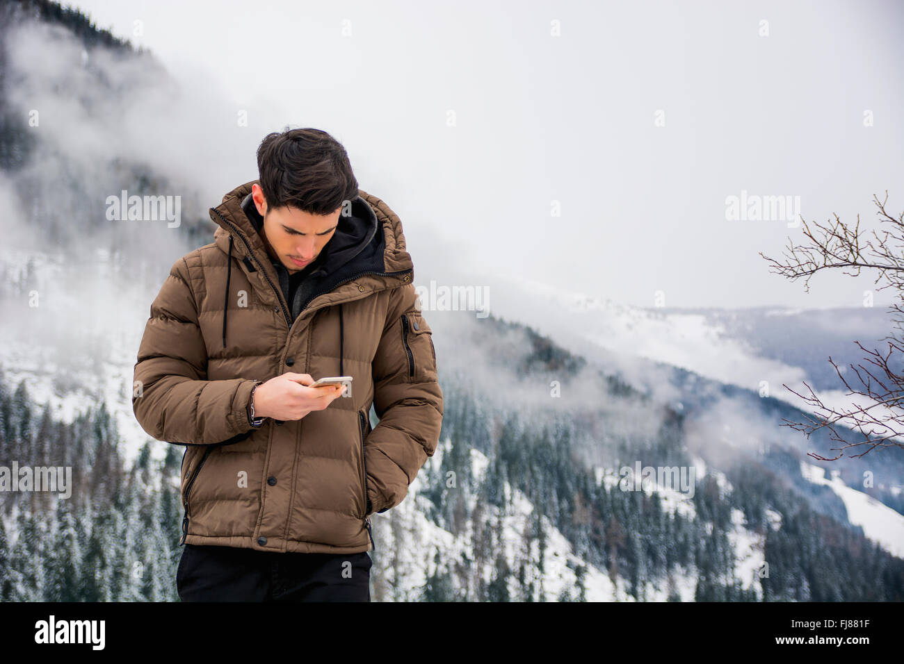 Dark haired handsome young man in winter outerwear using cell phone or smartphone, outdoor at mountain with snowy landscape behi Stock Photo