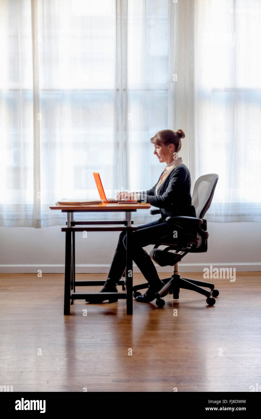 A woman sitting at a computer with window behind her Stock Photo