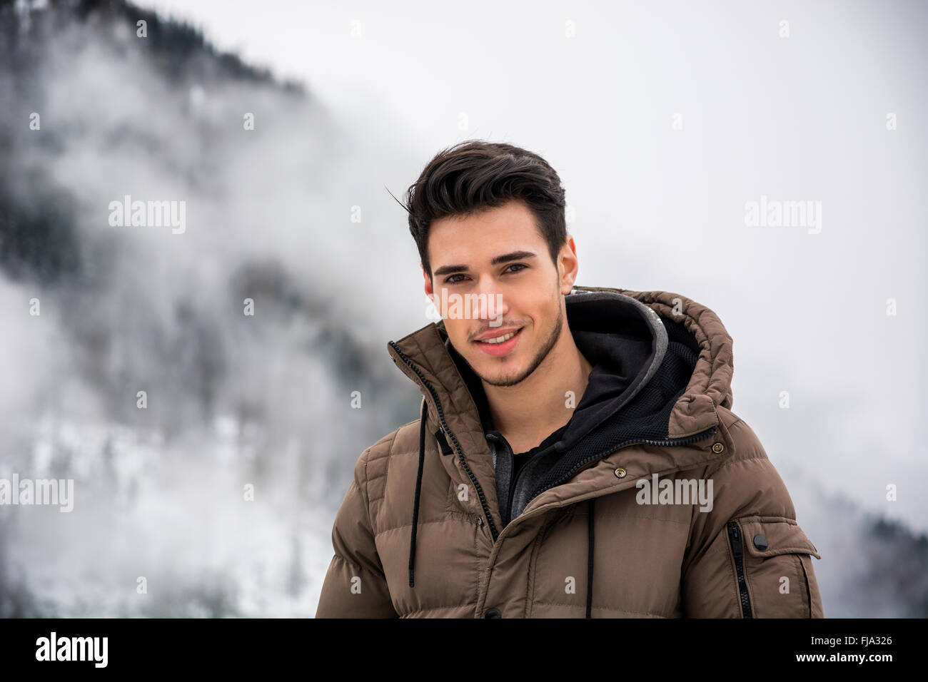 Handsome man in outerwear sitting while looking at camera. Snowy landscape on background Stock Photo