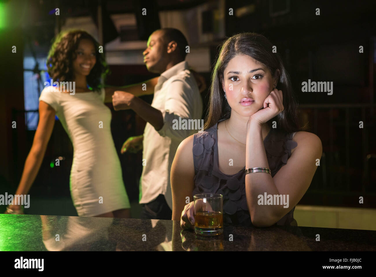 Unhappy woman sitting at bar counter and couple dancing behind her Stock Photo