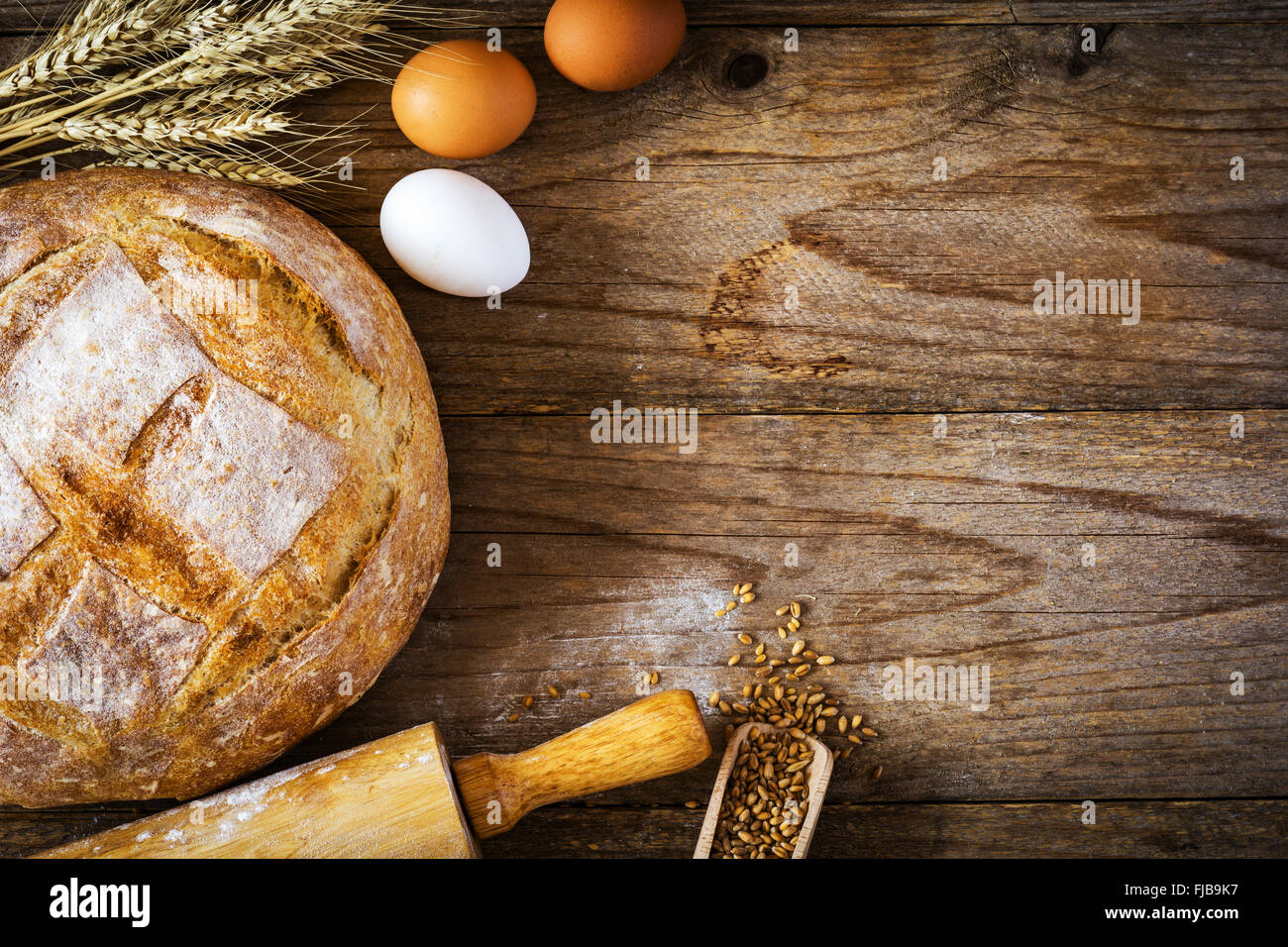 Baking bread food background: fresh round loaf of bread, fresh eggs, wheat, wheat ears and rolling-pin on wooden backdrop Stock Photo