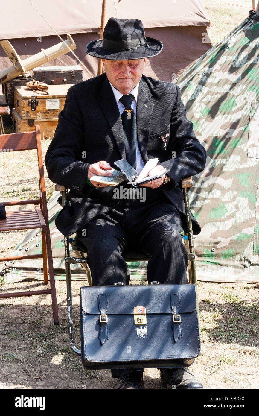 WW2 re-enactment at War and peace show. Senior male Gestapo official in black suit and hat, sitting outdoors, reading a book. Briefcase by feet. Stock Photo