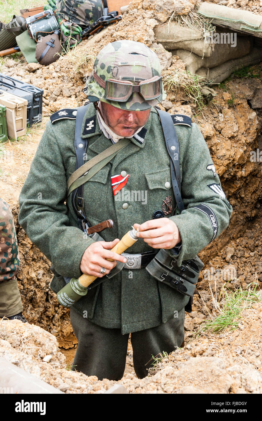 WW2 re-enactment. Looking down at German Waffen SS storm trooper in trench fusing a stick grenade, Stielhandgranate. Iron Cross ribbon pinned to chest Stock Photo