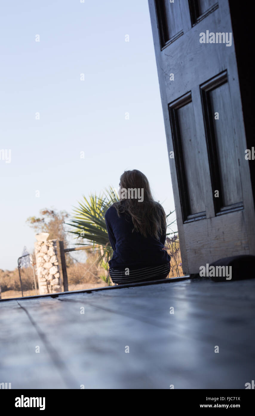 A young woman sits reflecting in the open doorway of an old house looking outside. Stock Photo