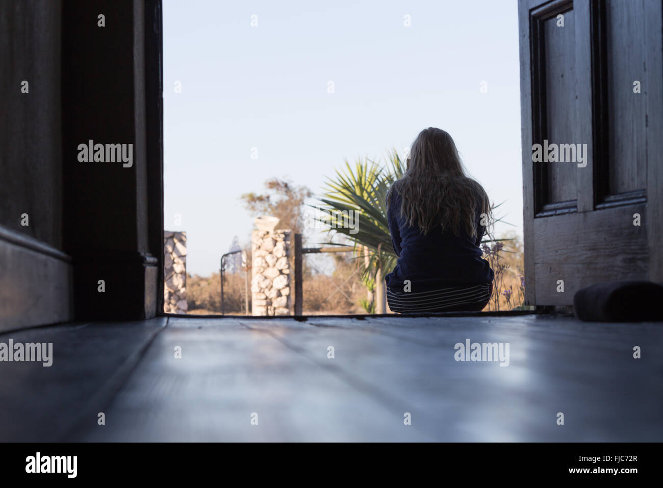 A young woman sits reflecting in the open doorway of an old house looking outside. Stock Photo