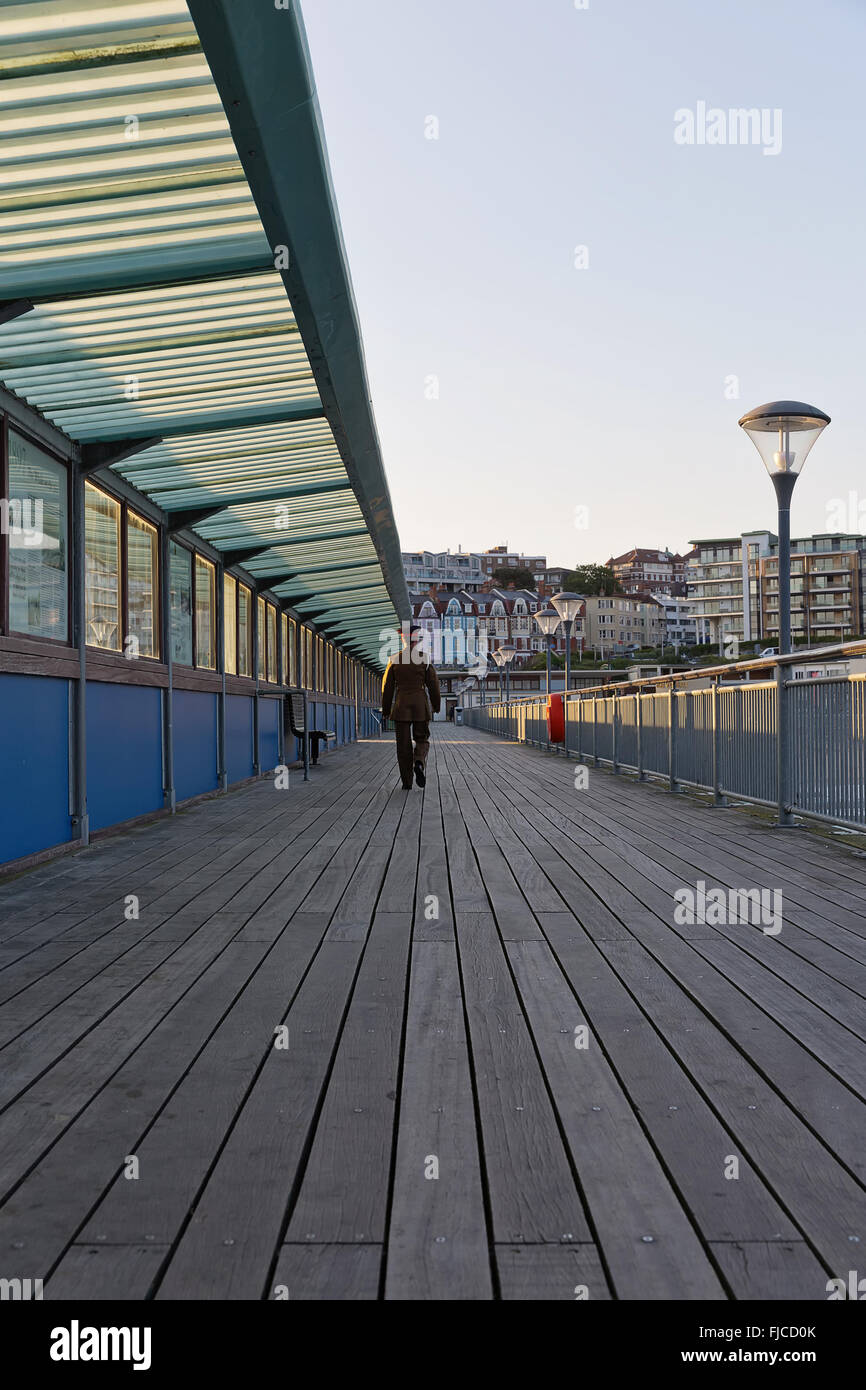 A guy in his army suit walking away from the camera, walking at the pier at sunset hour on a summer day with no clouds in the sk Stock Photo