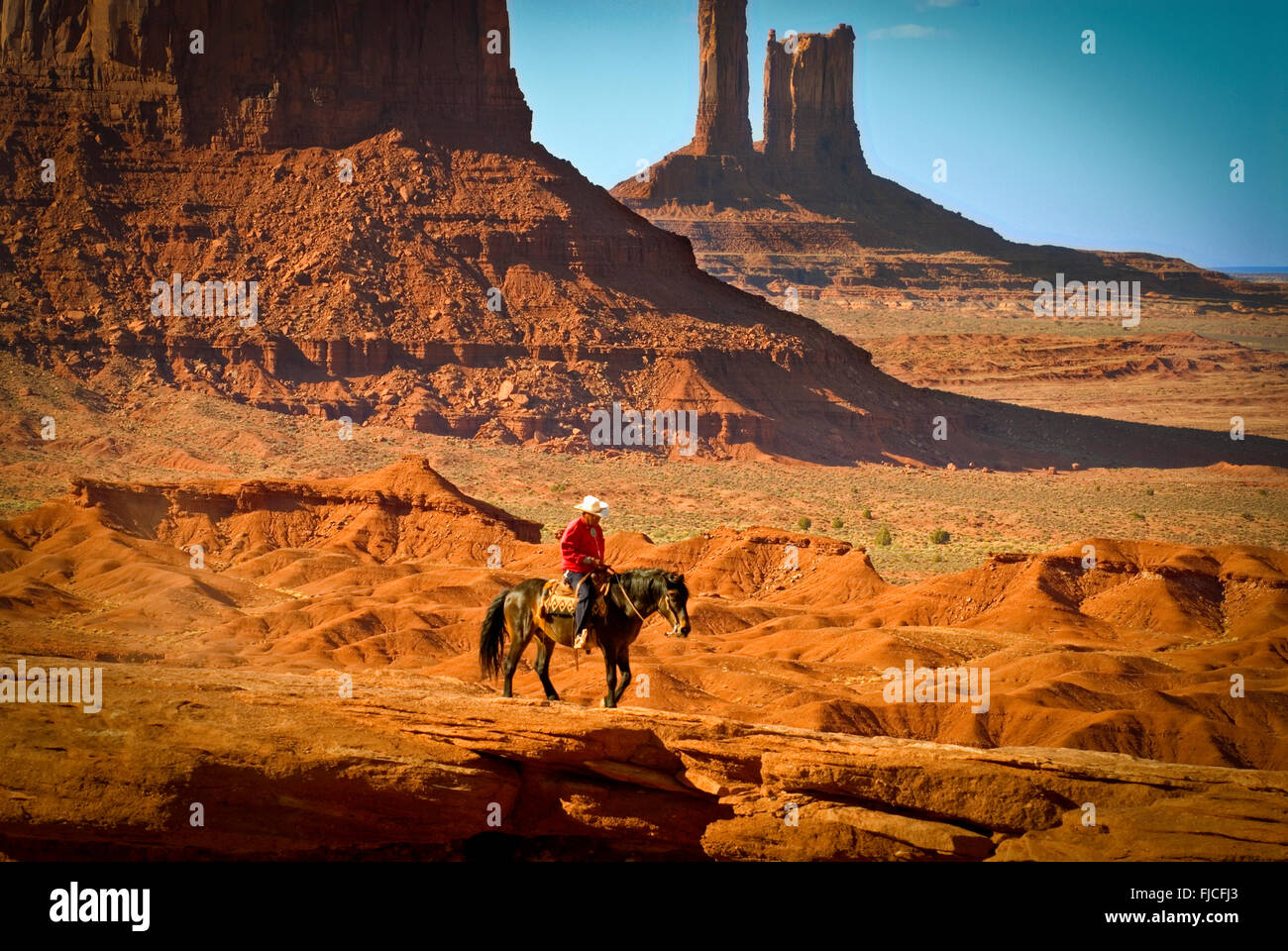 Navajo Indian cowboy riding his horse across colorful red sandstone plateau. Monument Valley Navajo Tribal Park, Arizona, USA Stock Photo