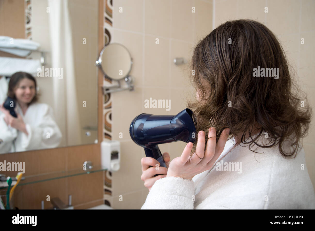 Portrait of young happy smiling woman wearing white bathrobe using hair dryer in bathroom, getting ready, drying her hair Stock Photo
