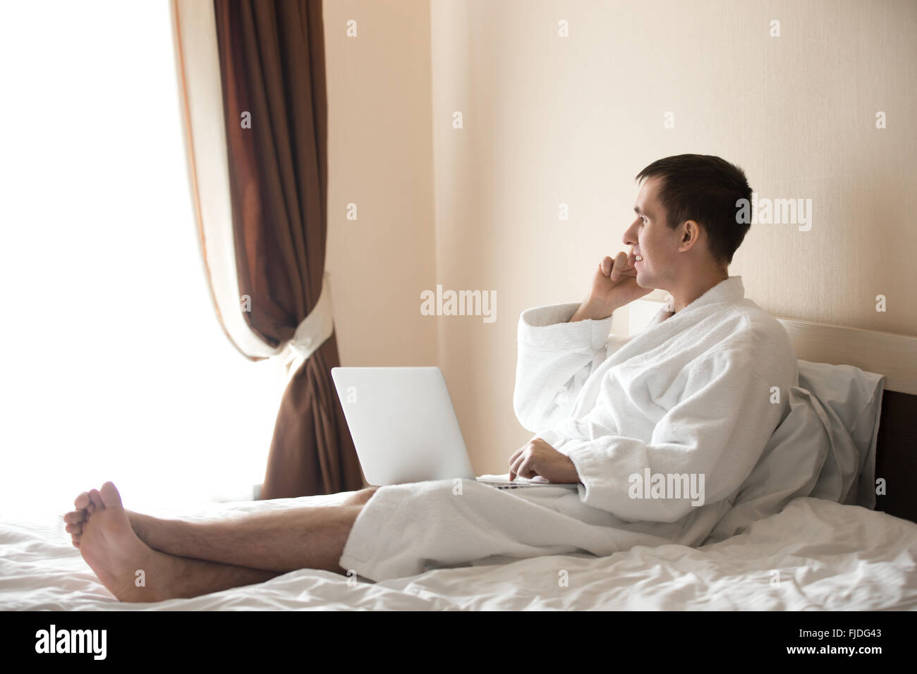 Portrait of attractive smiling young barefoot man wearing white bath robe sitting on bed with laptop, male model talking on cell Stock Photo