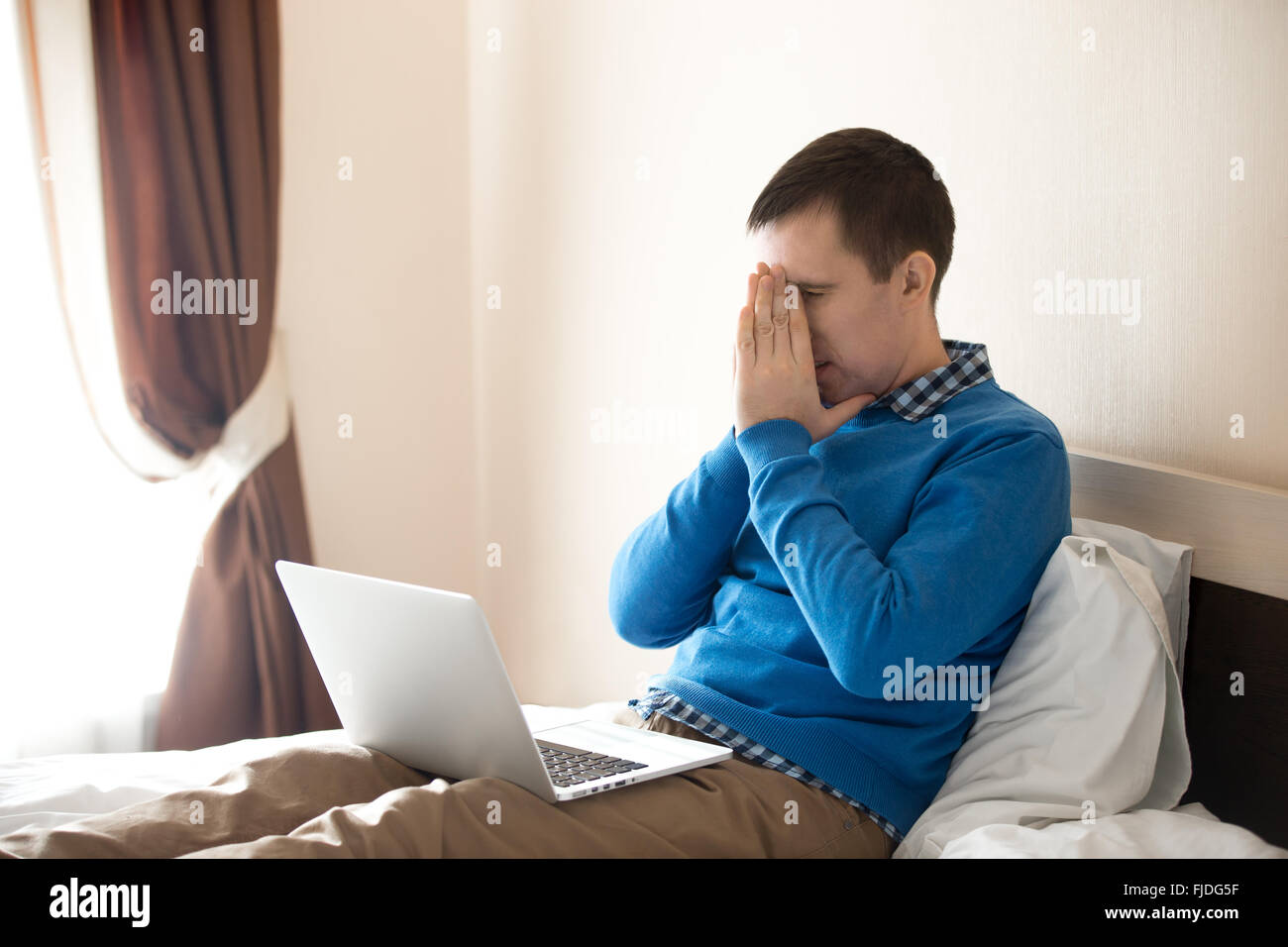Portrait of young stressed man sitting on bed with laptop wearing smart casual clothing, holding his head in hands Stock Photo