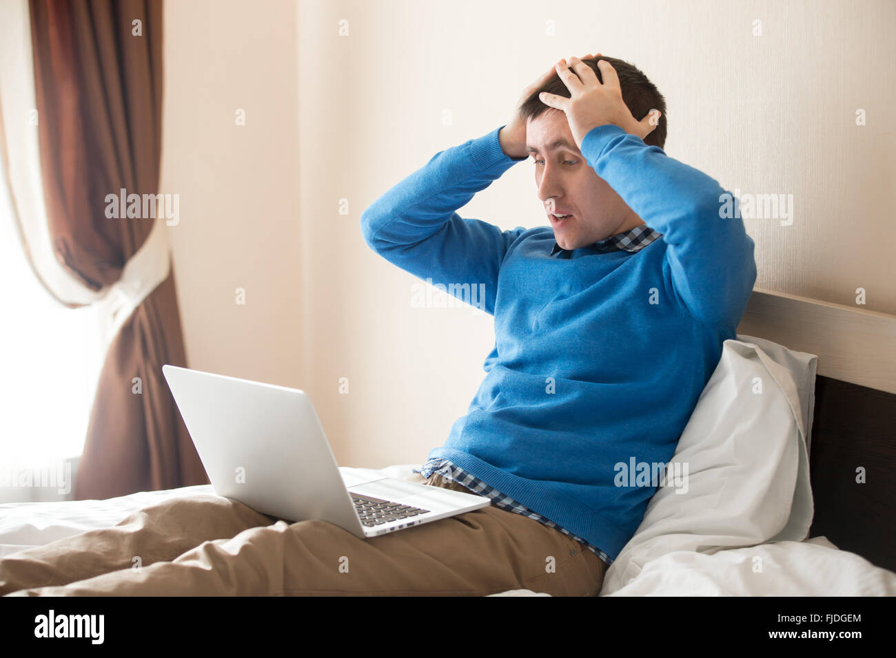 Portrait of young desperate man sitting on bed with laptop wearing smart casual clothing, holding his head in hands in dismay Stock Photo