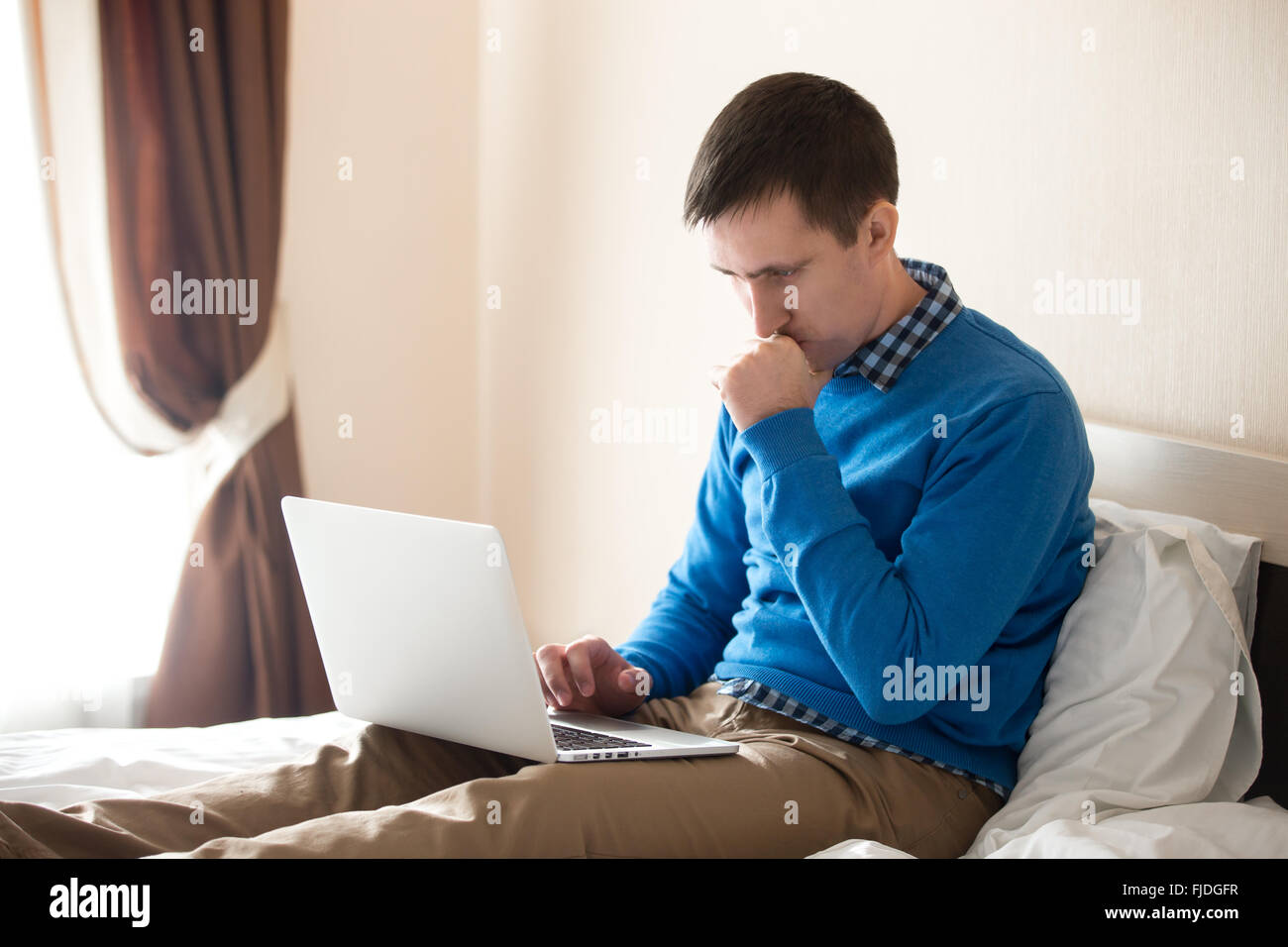 Portrait of young focused man sitting on bed with laptop wearing smart casual clothing, concentrated on work, male model Stock Photo