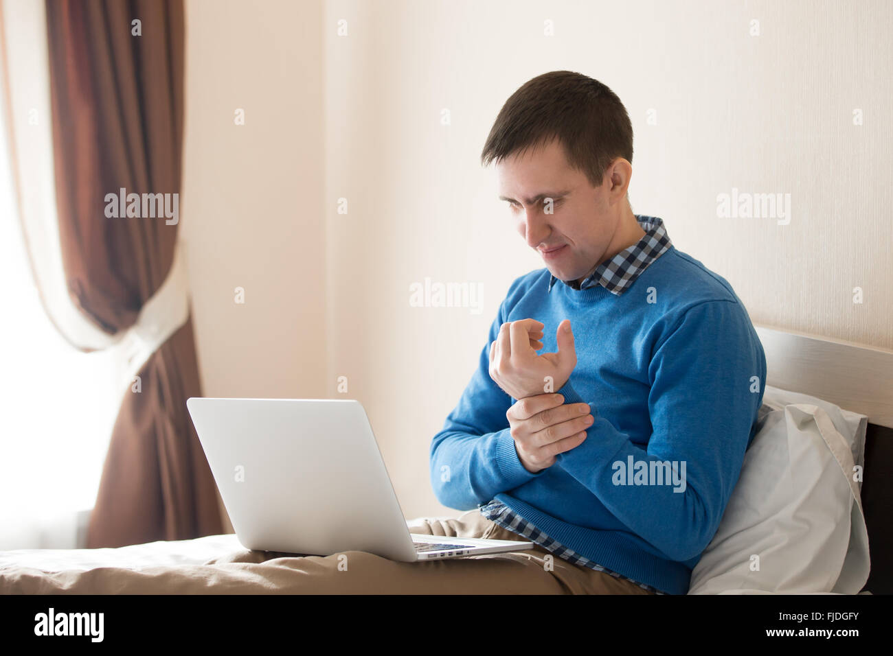 Portrait of young stressed man sitting on bed with laptop wearing smart casual clothing, touching his aching wrist with pained Stock Photo