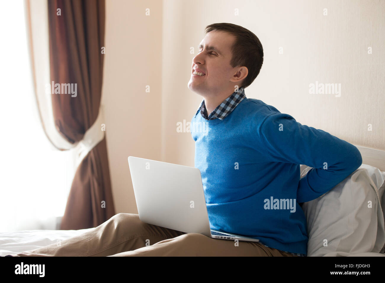 Portrait of young stressed man sitting on bed with laptop wearing smart casual clothing, feeling pain, touching his aching back Stock Photo