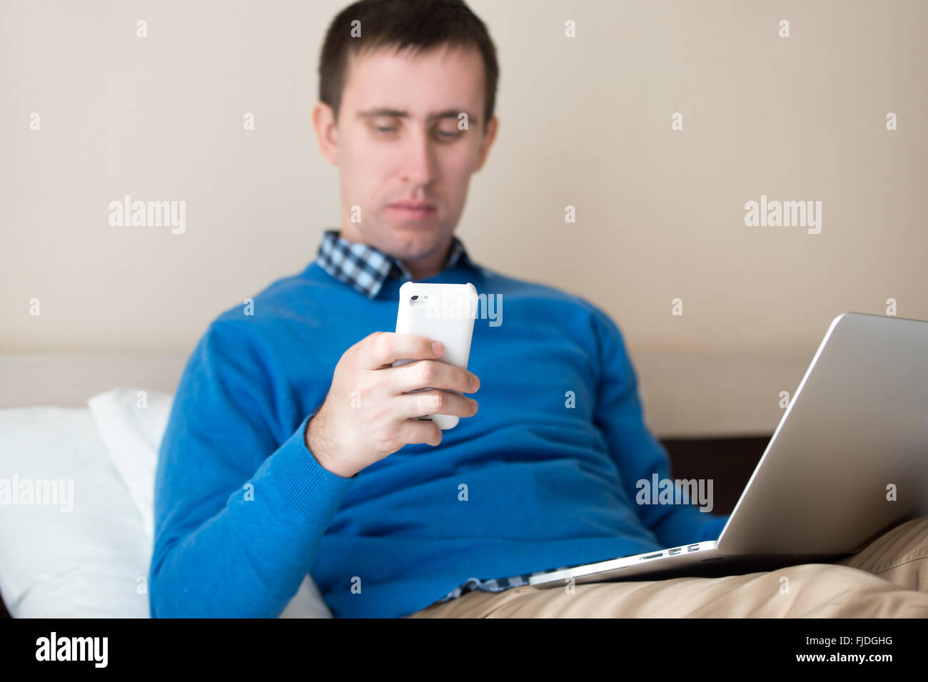 Portrait of attractive young man sitting on bed with laptop wearing smart casual clothing, using notebook and smartphone Stock Photo