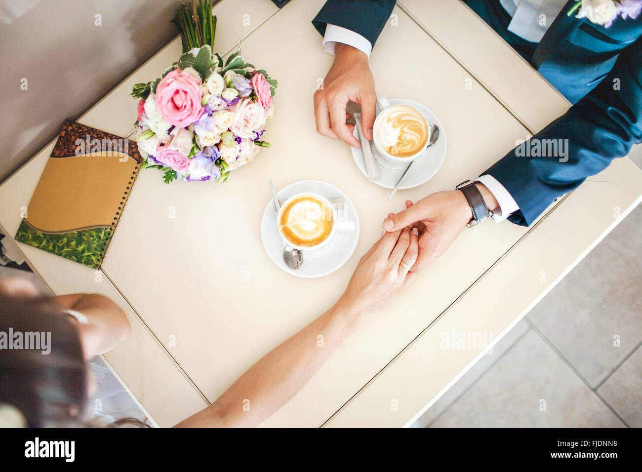 Wedding couple at cafe, top view. Man holds woman's hand, drinks espresso. Bride and groom coffee break dating gift, bouquet on table. Stock Photo