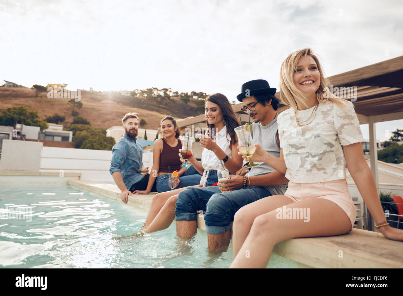 Multi-ethnic group of young people hanging out by swimming pool holding cocktails. Happy friends enjoying party by the pool. Stock Photo