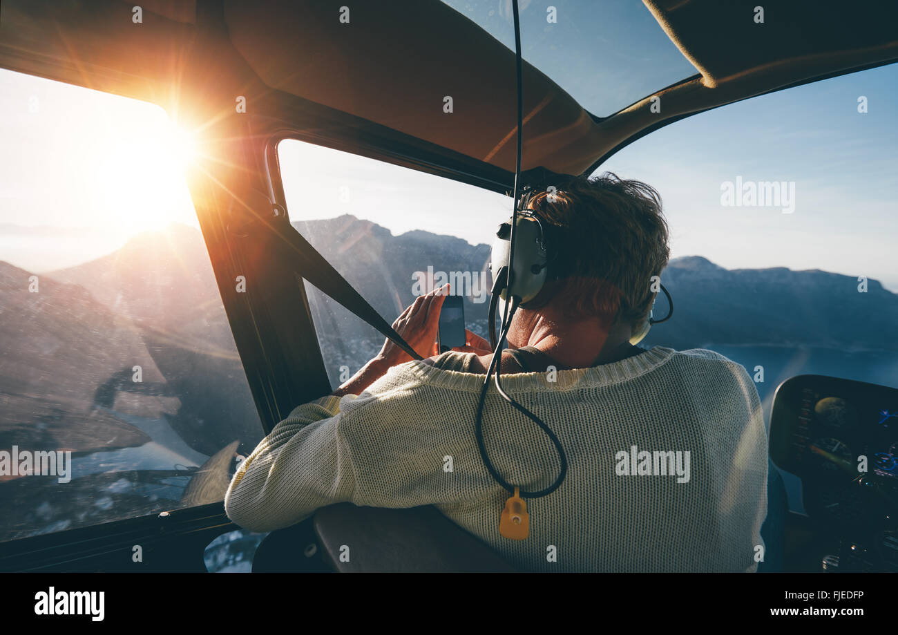 Rear view of female tourist on helicopter tour taking pictures while flying over mountains on a sunny day. Stock Photo