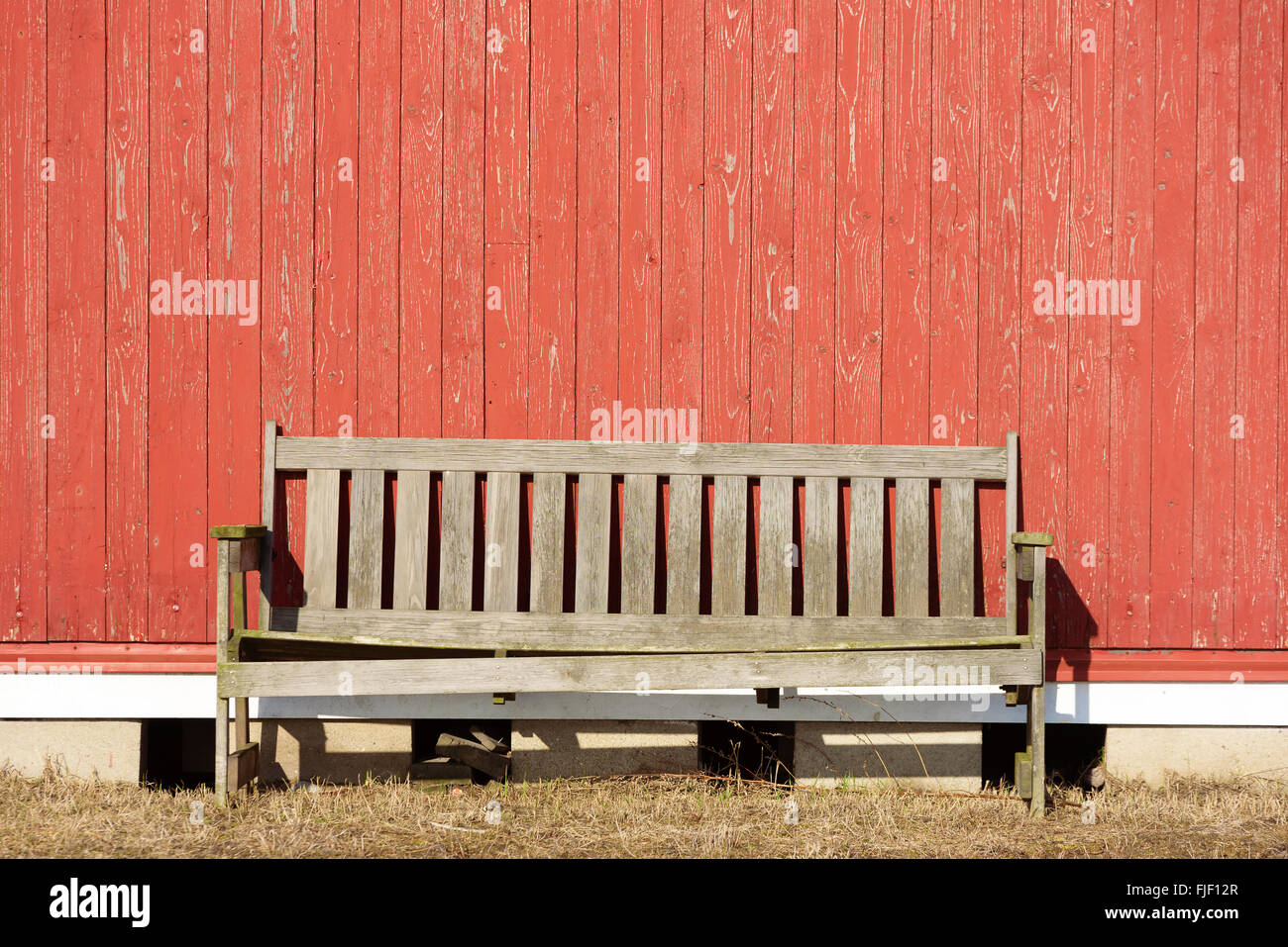 An old unpainted bench against a red wooden wall. Sun is shining at the empty outdoor furniture. Copy space on wall. Stock Photo