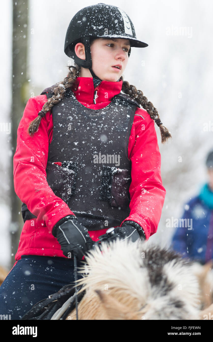 Girl with red jacket, black protective gear vest and helmet on a fjord horse at a riding lesson in snowfall looking aside Stock Photo