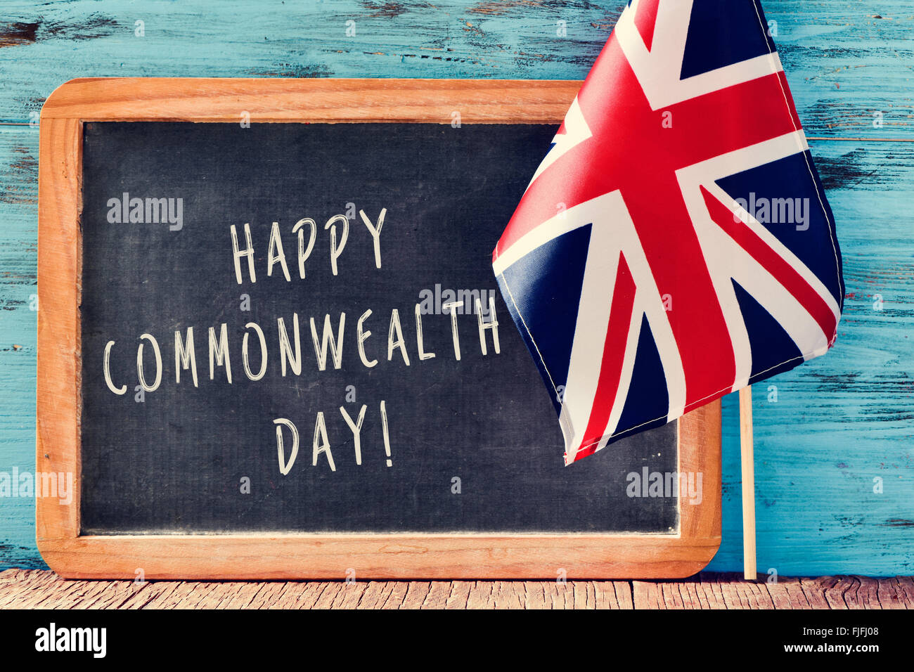 a chalkboard with the text happy commonwealth day written in it and the Union Flag, on a wooden surface, against a blue rustic w Stock Photo