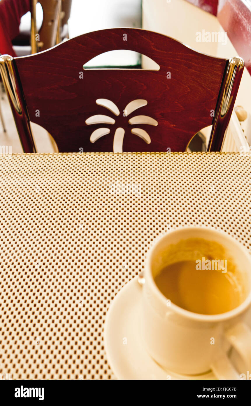 Wood-backed chair with palm tree motif is pulled up to a table with a dotted tablecloth design and a cup and saucer of coffee. Stock Photo