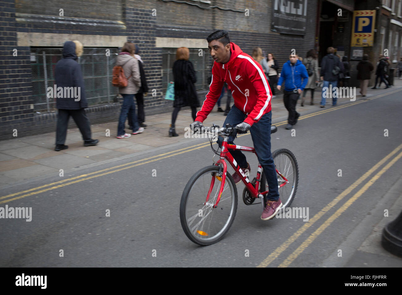 Young Asian man with a red bicycle and red tracksuit top cycling on Brick Lane in the East End of London UK. This area is particularly know for it s Indian Asian cuisine