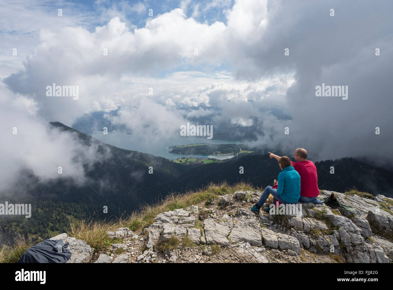Father pointing at Lake Walchensee beside his daughter sitting on a mountain summit watching cloud covered scenery Stock Photo